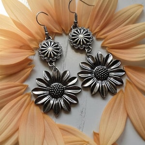 May include: Silver-coloured daisy earrings with a detailed floral design. The earrings hang from a simple hook and are set against a backdrop of soft orange flower petals, creating a delicate and natural aesthetic.