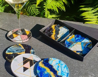 May include: Set of six coasters with abstract geometric and marble designs in shades of blue, black, white, gold, and pink. The coasters are displayed on a gray surface with a glass of white wine.
