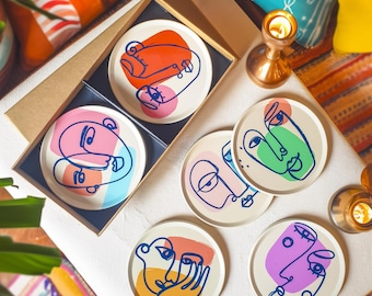 May include: Six colorful coasters with abstract faces drawn in blue lines. The coasters are round and have a white background. The faces are drawn in different colors, including pink, orange, green, and purple.