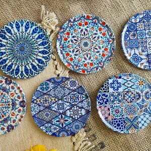 May include: Six round coasters with intricate blue, white, and red geometric patterns. The coasters are made of ceramic and have a glossy finish.