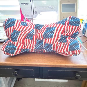 May include: Bone-shaped pillow featuring a vibrant American flag pattern. The pillow showcases the stars and stripes in red, white, and blue. It is placed on a wooden table, with a sewing machine in the background.
