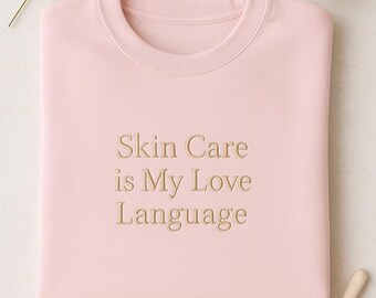 May include: A light pink sweatshirt with the embroidered text "Skin Care is My Love Language." The image includes a sprig of greenery, two small candles, and a cosmetic applicator.