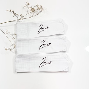 May include: Three white spa headbands with a waffle weave are displayed on a white surface. Each headband features the word "Zmar" embroidered in black script. A sprig of dried flowers is placed near the headbands.