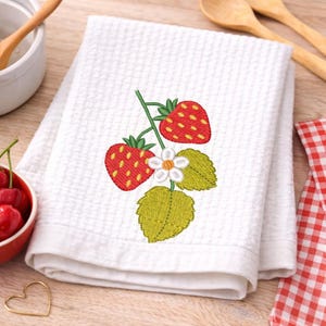 May include: White waffle-weave kitchen towel with an embroidered strawberry design. The design features two red strawberries with yellow seeds, a white flower, and green leaves. The towel is folded and appears clean.