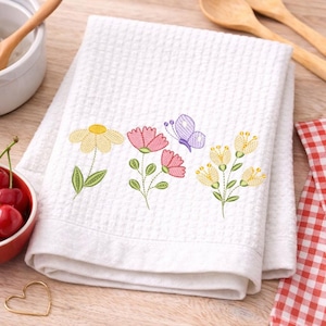 May include: White kitchen towel with a waffle weave texture. The towel is embroidered with colorful flowers and a butterfly. The flowers are yellow, pink, and the butterfly is purple. The towel is folded and placed on a wooden surface.