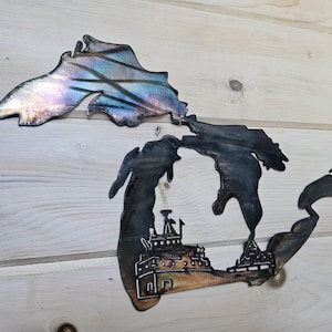 May include: Metal wall art depicting the outline of the Great Lakes. The upper portion has a shimmering, iridescent finish. The lower portion features a detailed illustration of a ship and buildings.