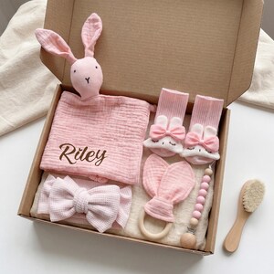 Può includere: Un set regalo per neonati rosa in una scatola di cartone. Il set include una coperta di sicurezza a forma di coniglietto con il nome "Riley", calzini con orecchie di coniglio, una fascia per capelli, un anello per la dentizione, una clip per ciuccio e una spazzola di legno.
