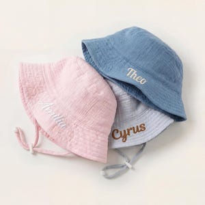 May include: Three personalized bucket hats in pastel colors: pink, blue, and gray. Each hat has a name embroidered on the front in a contrasting color: Amelia, Theo, and Cyrus. The hats have adjustable chin straps.