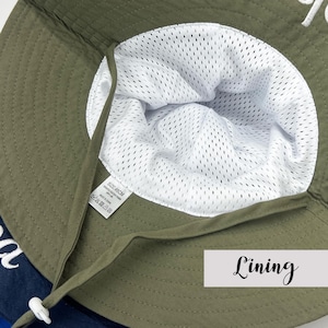 May include: An olive green bucket hat featuring a white mesh interior lining. The hat includes a matching olive green chin strap with a white plastic toggle. A small white tag inside displays the text SIZE 46CM, 100% POLYESTER FIBER, UPF 50+.