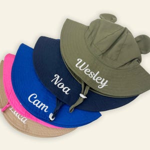 May include: A stack of five sun hats with ear-like accents on the crown and chin straps. The hats are colored olive green, navy blue, royal blue, hot pink, and tan. Each hat features a white embroidered name: Wesley, Noa, Cam, and Luca.