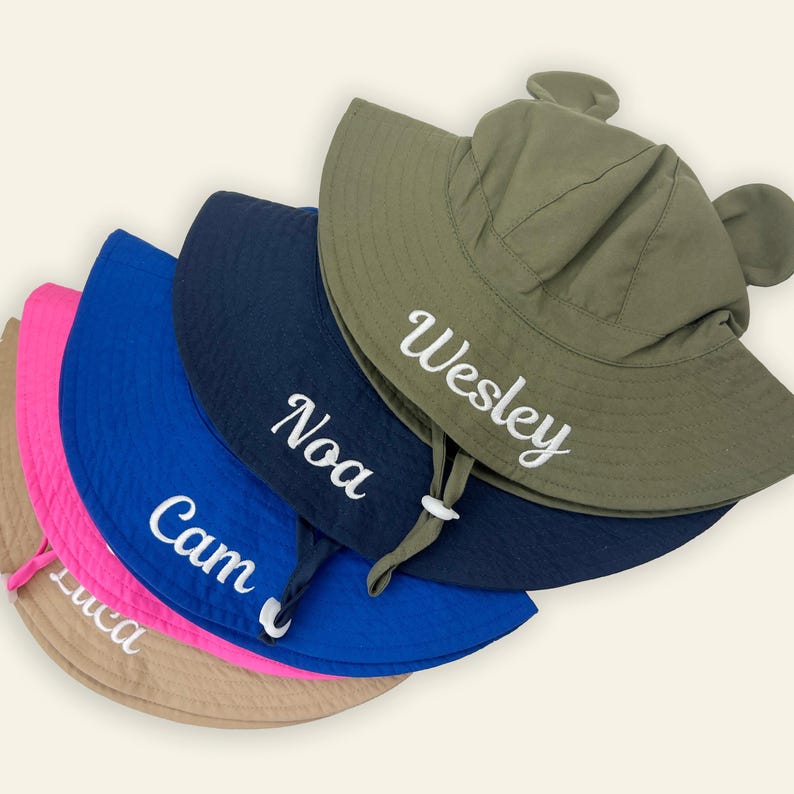 May include: A stack of five sun hats with ear-like accents on the crown and chin straps. The hats are colored olive green, navy blue, royal blue, hot pink, and tan. Each hat features a white embroidered name: Wesley, Noa, Cam, and Luca.