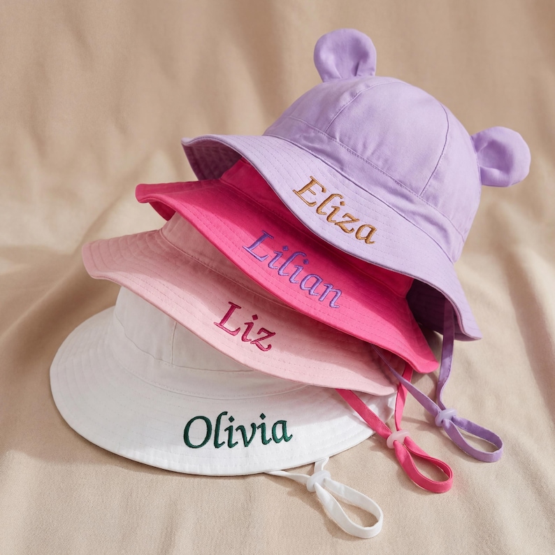 May include: A stack of four bucket hats in lavender, hot pink, light pink, and white. The lavender hat features bear ears. Each hat is embroidered with a name: Eliza, Lilian, Liz, and Olivia. Each hat includes an adjustable chin strap.