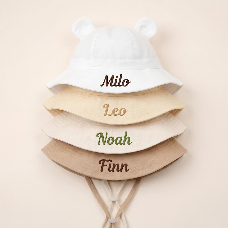 May include: A stack of four bucket hats with small bear ears on the top crown. The hats are white, cream, light beige, and tan. Each hat features embroidered names in cursive script: Milo in dark brown, Leo in tan, Noah in green, and Finn in brown.