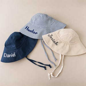 May include: Three personalized sun hats in navy, beige, and blue and white striped patterns. Each hat features a different name embroidered in a contrasting color: Daniel, Josiah, and Hunter. The hats have chin straps.
