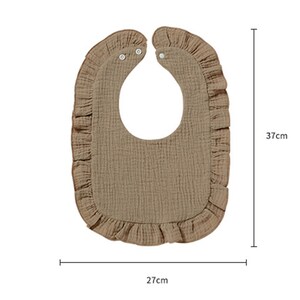 May include: A tan bib with a ruffled edge and two white snap closures at the neck. The bib features a textured fabric surface and measures 37 cm in height and 27 cm in width as indicated by the measurement lines and text printed on the image.