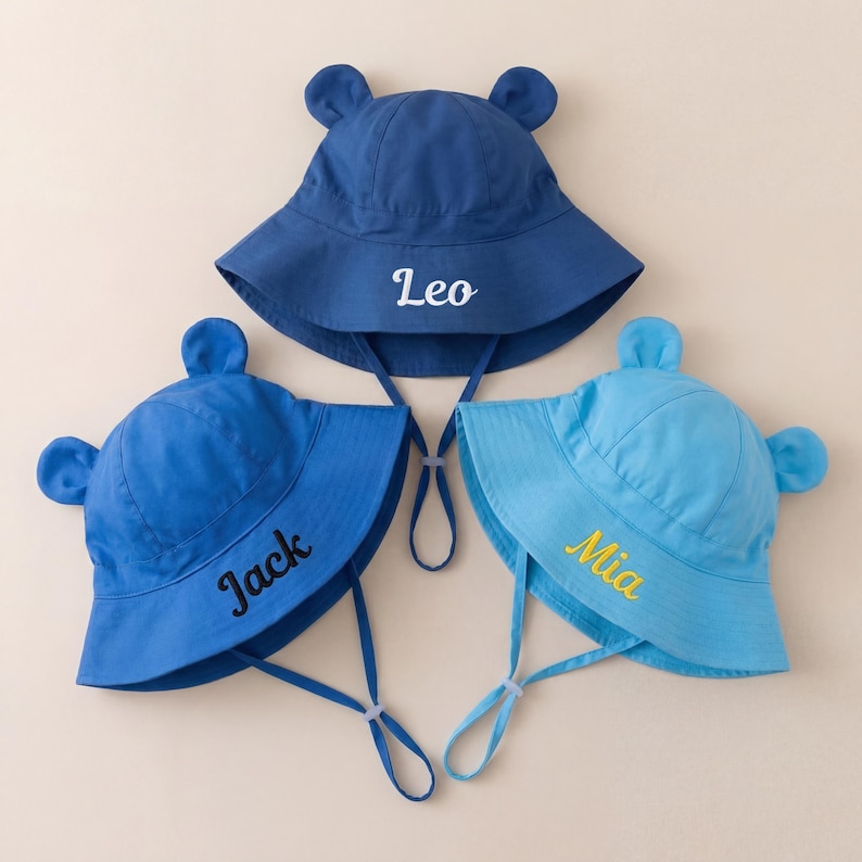 May include: Three bucket hats with bear ears and chin straps. The top hat is dark blue with white embroidered text reading Leo. The bottom left hat is blue with black embroidered text reading Jack. The bottom right hat is light blue with yellow embroidered text reading Mia.