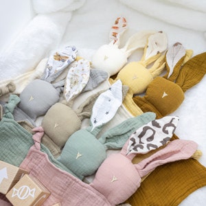May include: A set of plush bunny toys with different colored fabric ears and faces. The toys are arranged in a row on a pink and white blanket.