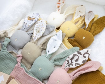 May include: A set of plush bunny toys with different colored fabric ears and faces. The toys are arranged in a row on a pink and white blanket.