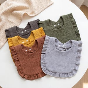 May include: A set of five ruffled bibs made of textured muslin fabric in shades of charcoal gray, mustard yellow, terracotta brown, olive green, and light gray. Each bib features a rounded shape with a ruffled edge and two white snap buttons.