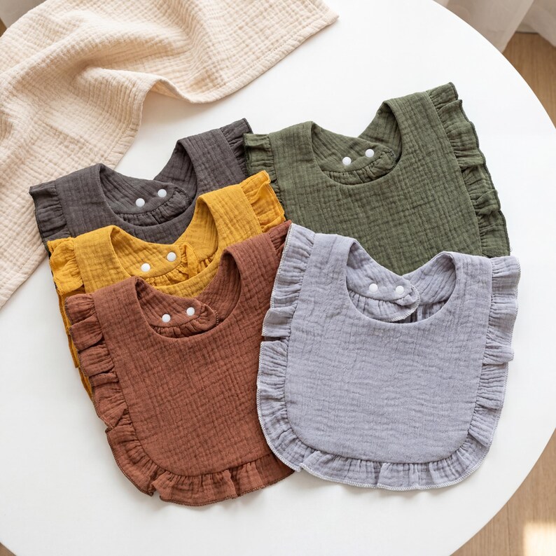 May include: A set of five ruffled bibs made of textured muslin fabric in shades of charcoal gray, mustard yellow, terracotta brown, olive green, and light gray. Each bib features a rounded shape with a ruffled edge and two white snap buttons.