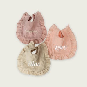 May include: Three ruffled baby bibs made of textured fabric in muted shades of dusty rose, peach, and beige. Each bib features white embroidered names in a cursive script: Henning on the rose bib, Riley on the peach bib, and Elias on the beige bib.
