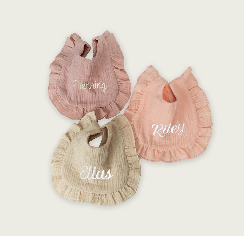 May include: Three ruffled baby bibs made of textured fabric in muted shades of dusty rose, peach, and beige. Each bib features white embroidered names in a cursive script: Henning on the rose bib, Riley on the peach bib, and Elias on the beige bib.
