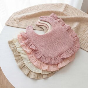 May include: A stack of four layered muslin bibs in shades of dusty rose, light pink, cream, and beige. Each bib features a rounded shape with a ruffled edge and a single white snap closure at the neck, crafted from a soft, textured fabric.