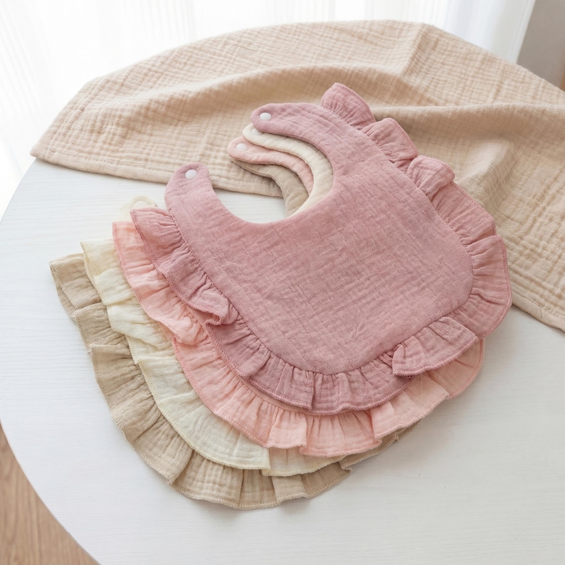 May include: A stack of four layered muslin bibs in shades of dusty rose, light pink, cream, and beige. Each bib features a rounded shape with a ruffled edge and a single white snap closure at the neck, crafted from a soft, textured fabric.