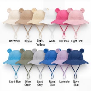 May include: A collection of twelve bucket hats with bear ears and chin straps, displayed in two rows. Colors include off-white, khaki, light yellow, white, hot pink, light pink, light blue, olive green, light grey, royal blue, lavender, and navy blue.