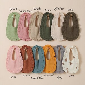 May include: A collection of twelve ruffled baby bibs in various colors including green, cameo pink, khaki, forest, off-white, olive, pink, bronze, muted blue, mustard, grey, and a white bib with a brown bear print, each featuring a button closure.