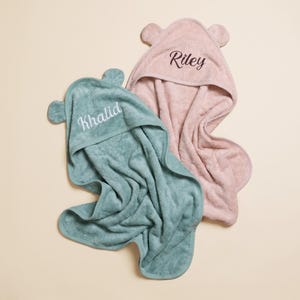 Peut inclure: Deux serviettes à capuche avec des oreilles d'ours sur les capuches. Une serviette est vert sauge avec le nom Khalid brodé en fil blanc. L'autre serviette est rose poudré avec le nom Riley brodé en fil marron foncé. Tissu doux et texturé.