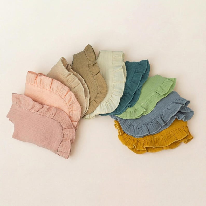 May include: A collection of ten semi-circular fabric bibs arranged in a fan shape, each featuring a ruffled edge. The colors include shades of dusty pink, peach, beige, tan, cream, teal, sage green, slate blue, and mustard yellow in a textured fabric.