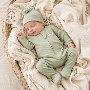 May include: A sage green ribbed long-sleeve footed sleeper and a matching knotted hat, both featuring the embroidered name Rex in white script. The garment includes a circular logo patch with the text Organic Cotton and a cotton plant icon.