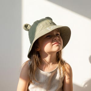 May include: An olive green bucket hat featuring two small rounded ears on the top and the name Leo embroidered in white thread on the front brim. The hat has a wide, downward-sloping brim and a structured crown, providing a simple and functional design.