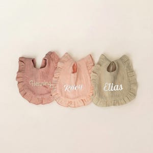 May include: Three muslin fabric bibs with ruffled edges in muted tones of dusty rose, light peach, and beige. Each bib features white embroidered text in a cursive script, displaying the names Henning, Riley, and Elias respectively.