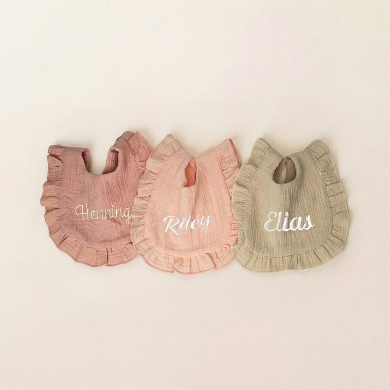 May include: Three muslin fabric bibs with ruffled edges in muted tones of dusty rose, light peach, and beige. Each bib features white embroidered text in a cursive script, displaying the names Henning, Riley, and Elias respectively.