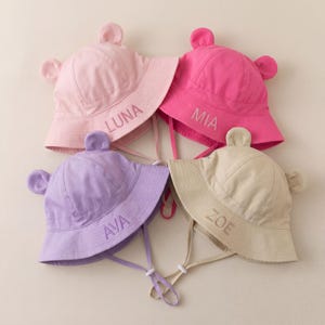 May include: Four bucket hats with bear ears, each featuring an adjustable chin strap. The hats are light pink with the name LUNA, hot pink with MIA, light purple with AVA, and beige with ZOE embroidered in matching thread on the front brim.