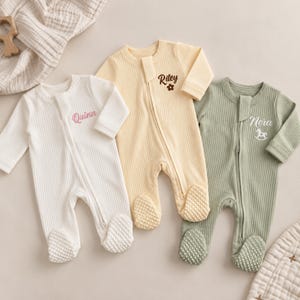 May include: Three long-sleeved, ribbed footed sleepsuits in white, pale yellow, and sage green. Each features a front zip fastening and a personalised name embroidered on the chest: Quinn in pink, Riley with a flower, and Nora with a rocking horse.