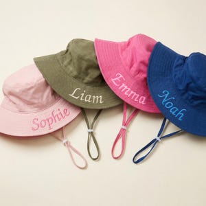 May include: Five colourful bucket hats with embroidered names. The hats are in pink, green, and blue, each with a matching chin strap. The names "Sophie", "Liam", "Emma", and "Noah" are embroidered in a contrasting colour. Ideal for children.