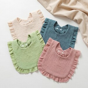 May include: Four muslin baby bibs with ruffled edges, each featuring a different solid color: beige, sage green, dusty rose, and teal blue. Each bib has a rounded shape and includes two white snap buttons at the neck for secure closure.
