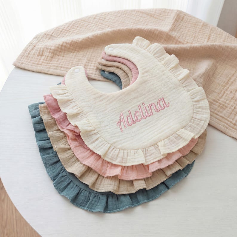 May include: A stack of four ruffled muslin bibs in cream, dusty pink, beige, and muted blue. The top cream bib features the name Adelina embroidered in pink thread. Each bib has a rounded shape with a snap closure and a gathered fabric ruffle.