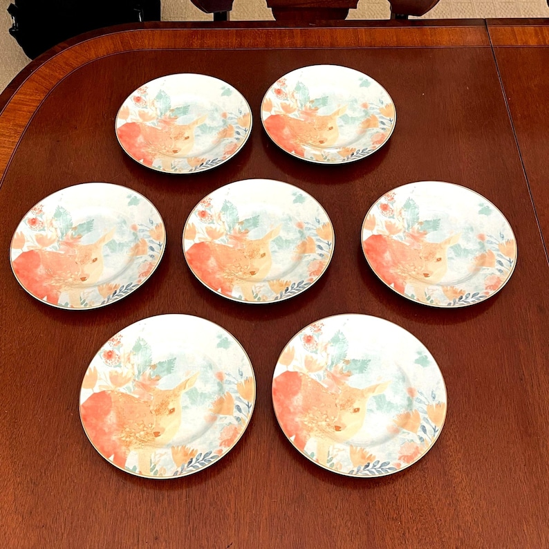 Puede incluir: Siete platos de cer&aacute;mica blanca con un dise&ntilde;o decorativo que presenta una vaca naranja claro, rodeada de elementos florales en tonos naranja, verde y azul. Los platos tienen un borde dorado y est&aacute;n dispuestos sobre una superficie de madera marr&oacute;n oscuro.