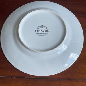Puede incluir: Un plato de cer&aacute;mica blanco con un borde ancho, visto desde la parte inferior. El centro presenta el texto "PATINA VIE HISTORY & STYLE COLLIDE" en un emblema decorativo. El plato est&aacute; sobre una superficie de madera oscura.