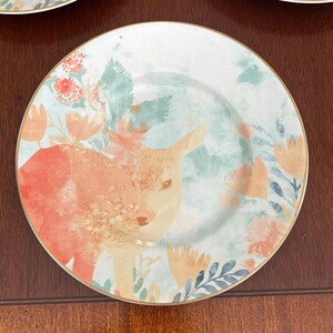 Puede incluir: Un plato redondo blanco con borde dorado, con un dise&ntilde;o de acuarela de un cervatillo y elementos florales en tonos naranja, rosa y azul. El plato tiene una est&eacute;tica delicada y art&iacute;stica, adecuado para servir alimentos.