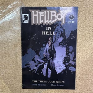 May include: A Hellboy In Hell issue 5 comic book with a dark blue and black cover featuring the title, the number 5, and the text The Three Gold Whips, Mike Mignola, and Dave Stewart. The illustration depicts Hellboy standing in a graveyard.