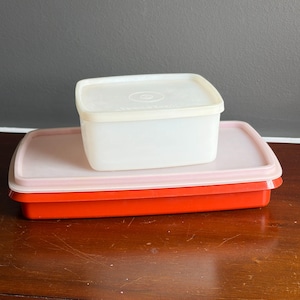 Puede incluir: Tres recipientes de plástico para guardar alimentos apilados. El recipiente inferior es rectangular y de color rojo brillante. El recipiente del medio es de color rosa claro, y el superior es cuadrado y blanco.