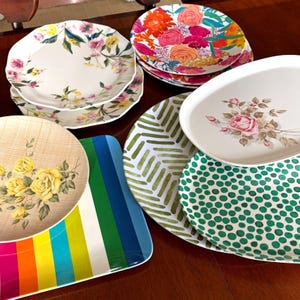 Op de afbeelding: Een verzameling decoratieve borden en serveerschalen met diverse bloemen- en geometrische patronen. De borden hebben ontwerpen met roze, gele en oranje bloemen. Serveerschalen omvatten een gestreept ontwerp en een groen stippenpatroon.