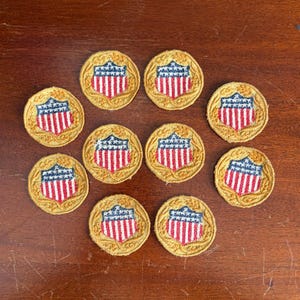 May include: A collection of ten circular embroidered patches. Each patch features a shield design with a blue top, red and white stripes, and a gold border. The patches are arranged on a wooden surface.