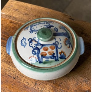 May include: A round ceramic casserole dish with a lid, featuring a hand-painted blue and orange cow motif on a cream background. The lid has a green knob, and the base is accented with a green rim and two small blue side handles for easy lifting.