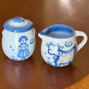 Puede incluir: Una taza de cerámica blanca con asa negra y el texto "But First, Coffee" en negrita negra. La taza se encuentra sobre un fondo borroso, sugiriendo un ambiente acogedor.